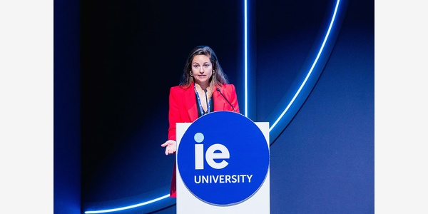 A speaker in a red outfit stands at a podium with the IE University logo.