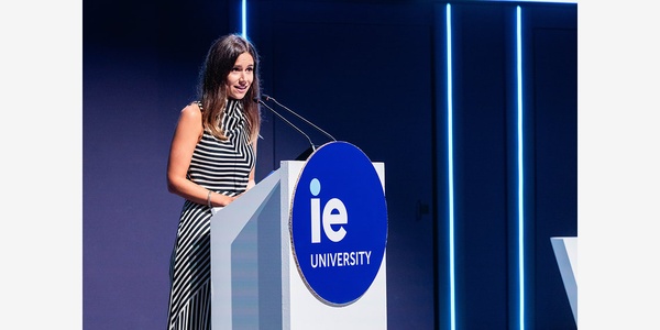 A woman is speaking at a podium with the logo of IE University in a modern setting.