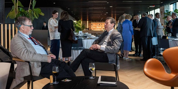 Two men are seated at a table in a modern restaurant, engaged in conversation while others socialize in the background.