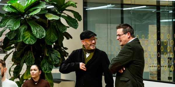 Two men in a casual business setting, engaging in a cheerful conversation near a large potted plant.