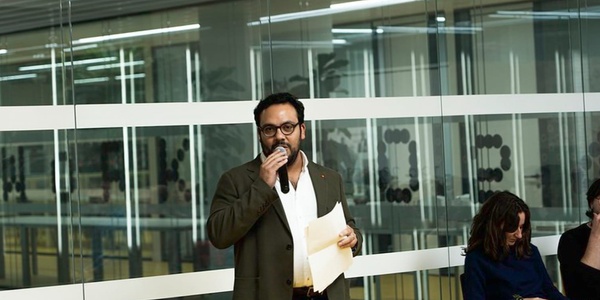 A man with glasses and a beard is speaking into a microphone in an office setting with other seated people listening.