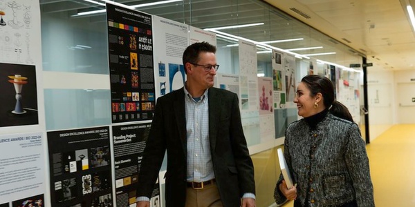 Two people, a man and a woman, walking and talking in a corridor with informational posters on the walls.