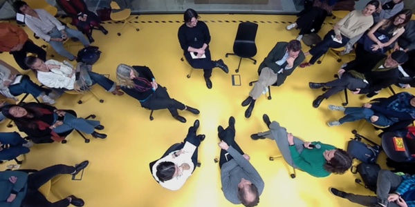 An overhead view of a group of people sitting and standing on a yellow floor, some conversing and others using smartphones.