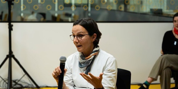 A woman is speaking into a microphone while gesturing with her hands during an indoor event.
