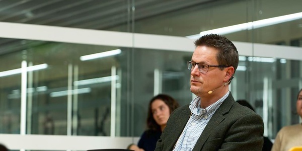 A man wearing glasses and a gray blazer attends a meeting in a modern office setting.