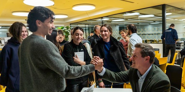 Two men shaking hands in a bustling office environment, surrounded by bystanders.