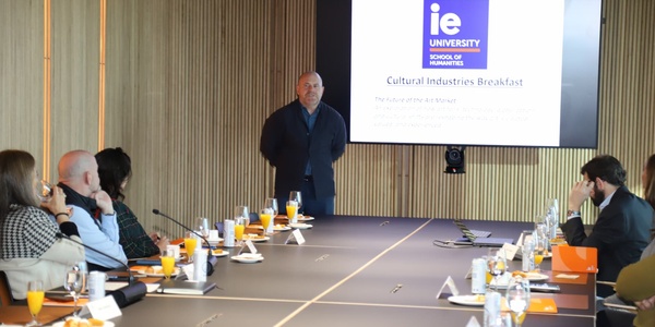 A speaker presents at a Cultural Industries Breakfast event attended by various individuals seated around a conference table.