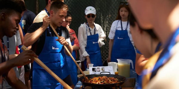 People in blue aprons are cooking and stirring a large paella outdoors.