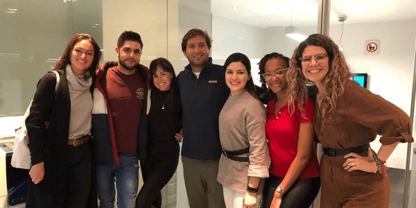 A group of seven people smiling and posing together indoors, with a sign in the background indicating no smoking.