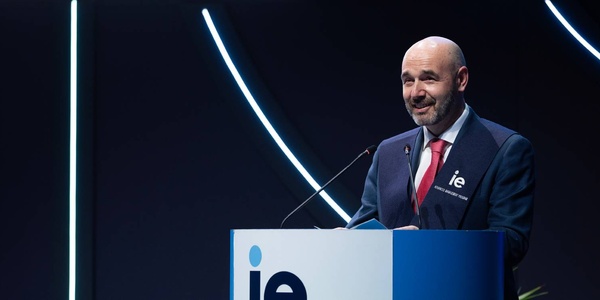 A man speaking at a podium with the letters 'IE' visible, surrounded by modern lighting design.