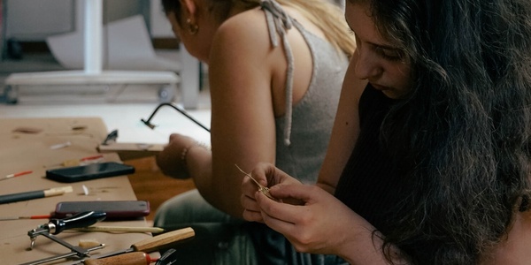 Two women are focused on crafting jewelry at a workshop table cluttered with tools and materials.