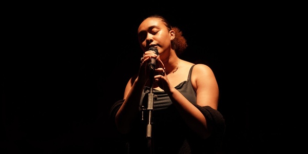 A woman singing into a microphone on a dark stage, illuminated by a soft spotlight.