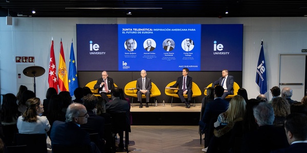 Four men are seated on stage at a panel discussion in a modern conference setting with logos of IE University and monitor displays in the background.