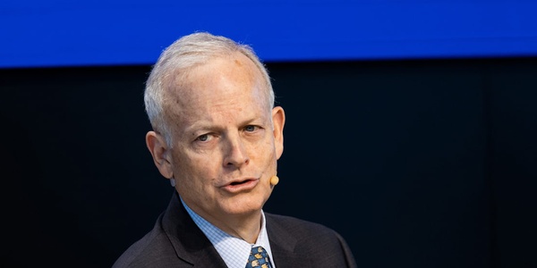 A mature man with gray hair, wearing a suit and tie, is speaking at a podium with a blue and yellow background.