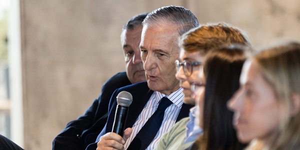 A senior man speaks into a microphone at a panel discussion while seated with other panelists.