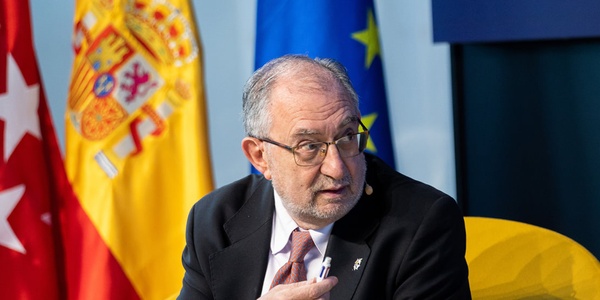 A man in a suit gestures while speaking, with the flags of Spain, the European Union, and Madrid in the background.
