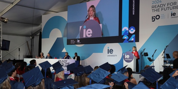 A graduation ceremony at IE University with students in blue gowns and a speaker on stage.