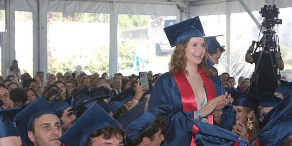 A young woman in a graduation cap and gown stands smiling and applauding among fellow graduates at a commencement ceremony.
