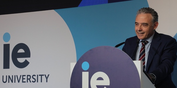 A man in a suit speaking at a podium with the IE University logo on it.