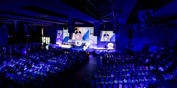 A large audience at a conference with a speaker on stage and multiple screens displaying the event.