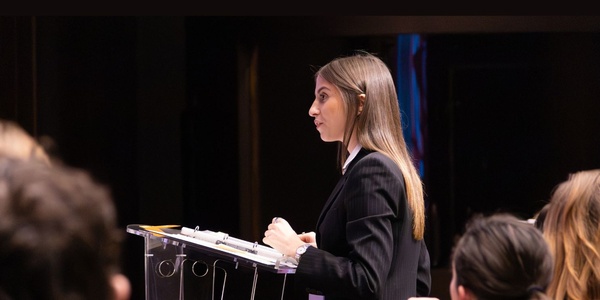 A young woman is delivering a speech at a podium in front of an audience.
