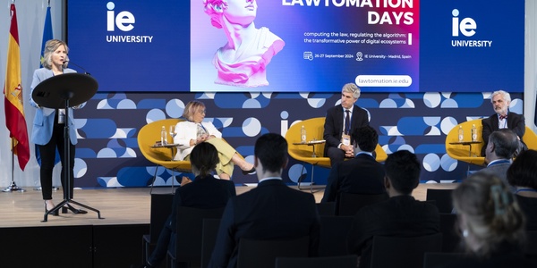 A woman speaks at a podium during a panel discussion at the Lawtomation Days event, with other panelists and an audience in attendance.