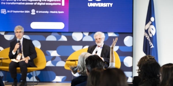 Two men sitting in front of an audience at a university event, with one speaking and the other listening.