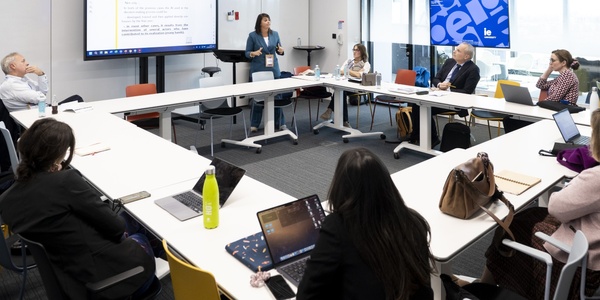 A woman stands presenting to a group of eight people seated around a conference table in a modern office.