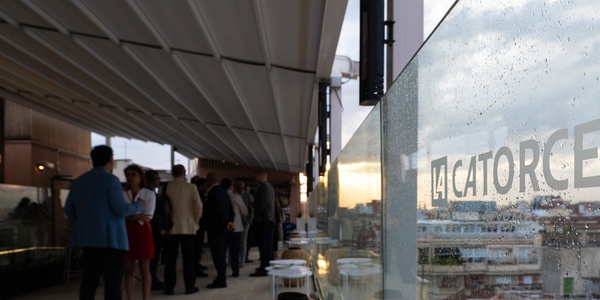 The image depicts a social gathering on a rooftop terrace with a view, featuring people chatting and glass barriers with reflections.