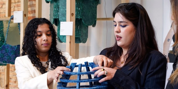 Two women, one holding a blue woven piece, are discussing or demonstrating a textile craft in a workshop with various fabrics displayed in the background.