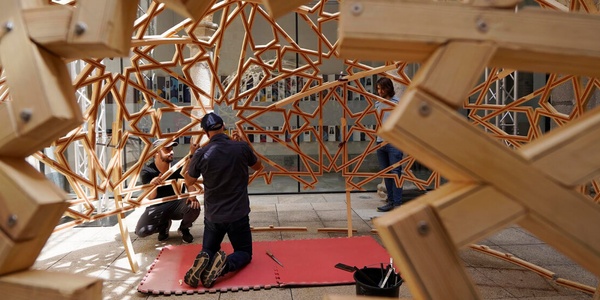 Two people are assembling a large geometric wooden structure outdoors.