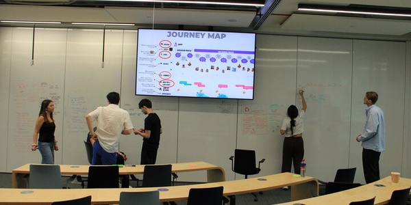 A group of people collaborating in a classroom setting while referring to a journey map displayed on a screen.