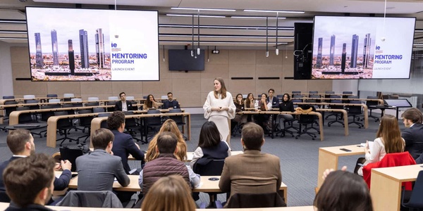 A woman stands presenting at the front of a conference room filled with seated participants, with two large screens displaying 'MENTORING PROGRAM'.