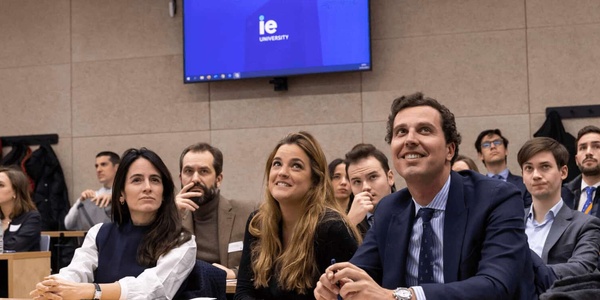 A group of people sitting in a conference room looking attentively towards a presentation screen displaying 'IE University'.