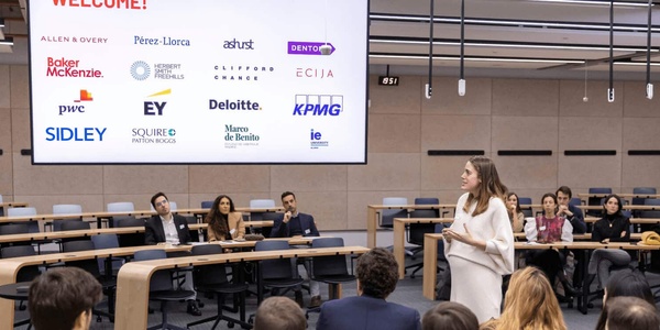 A woman stands speaking at the front of a lecture hall filled with seated attendees, with several professional service firm logos displayed behind her.