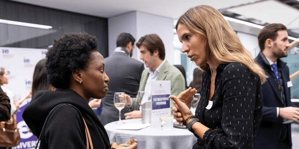 Two women engage in conversation at a professional networking event, with other attendees in the background.