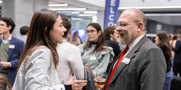 A young woman and an older man engaging in a conversation at a networking event, with other attendees in the background.