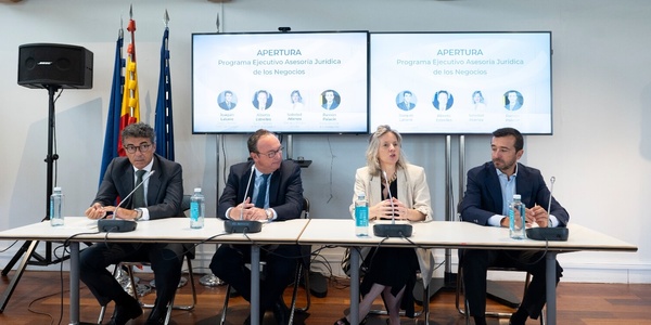 Four people sitting at a panel discussion table in a conference room, with projectors displaying the word 'APERTURA' in the background.