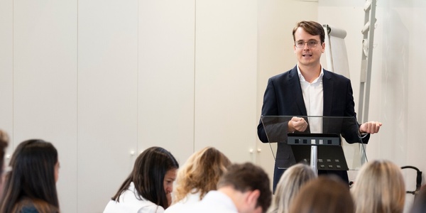 A young man giving a presentation at a podium in front of an audience of seated women.