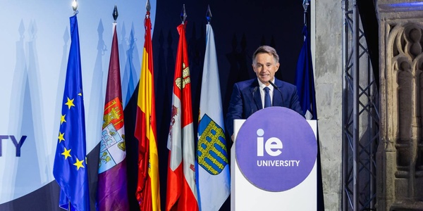 A speaker stands at a podium with various flags in the background during an event for IE University.