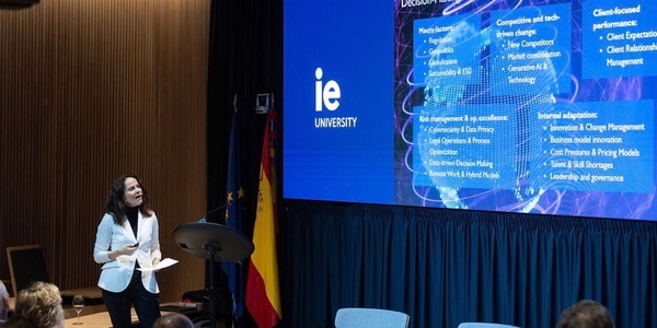 A speaker presents information at an event, with a digital screen displaying content related to IE University.