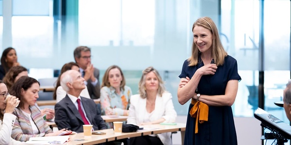 A woman stands smiling at a podium in a seminar room while a group of attentive adults are seated listening to her.
