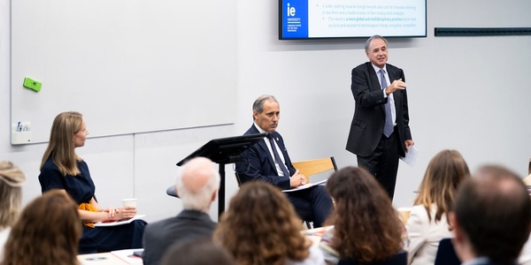 A man speaks at a podium in a lecture hall with attentive audience and two other participants seated.