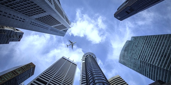 A plane flies above skyscrapers under a partly cloudy sky.