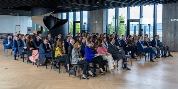 A group of people seated in rows at an indoor event, facing a focal point not visible in the image.