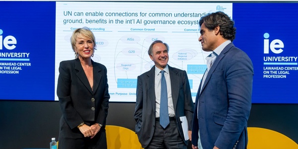 Three professionals are smiling and interacting in a seminar environment, with a backdrop displaying a presentation about legal frameworks and IE University's branding.