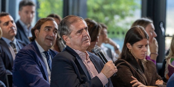 An older man holding a microphone speaks at a conference, surrounded by other attendees listening attentively.