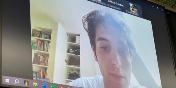 A young man appears on a video call on a computer screen, with a bookshelf in the background.