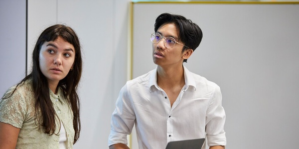 Two young individuals appear to be engaged in a discussion while standing near a laptop.