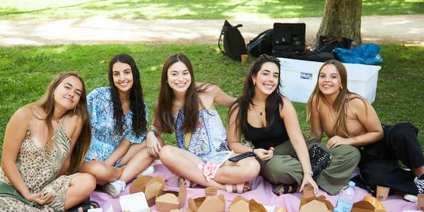 Five young women enjoying a picnic in the park with various food items spread out on a pink blanket.